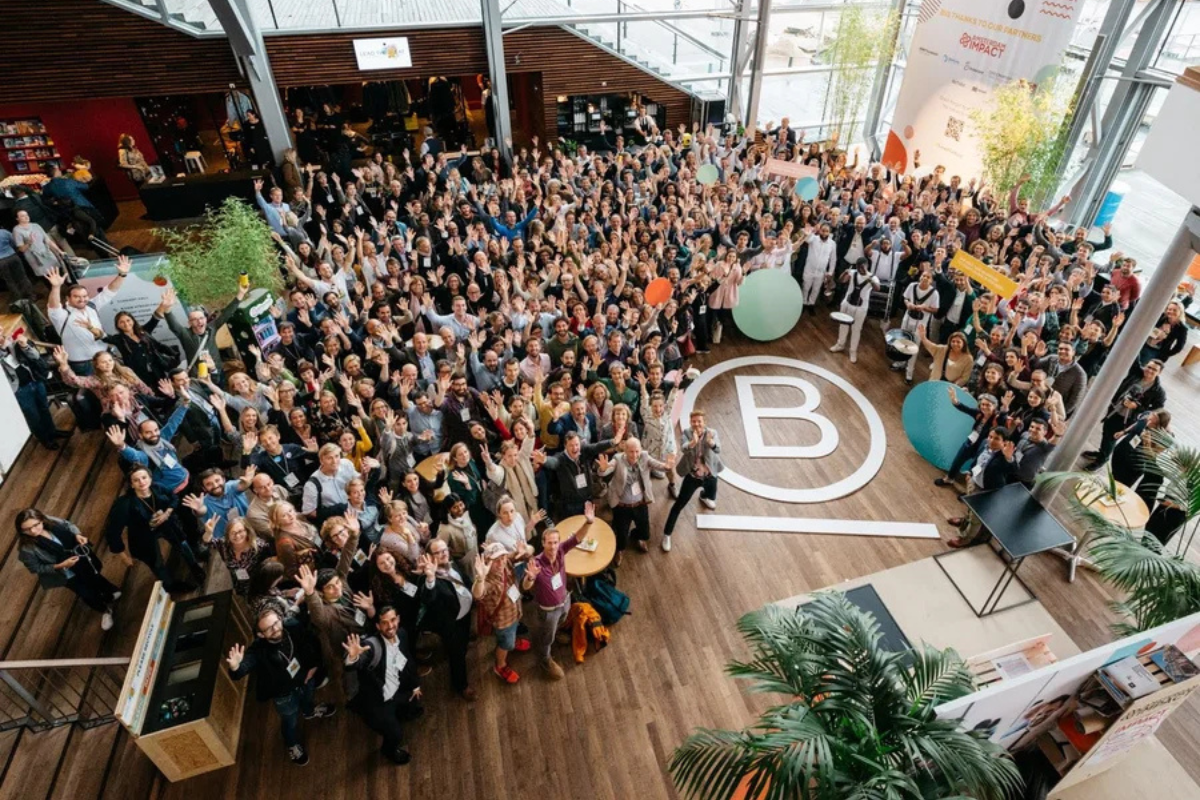 A large group of people gathered in a bright, modern space, celebrating a Certified B Corporation (B Corp) event. The floor features a prominent B Corp logo, symbolising the collective commitment to social and environmental impact